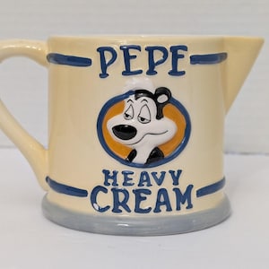 May include: Cream-colored ceramic creamer with a handle and spout. The creamer features a cartoon character illustration in a blue circle, with the words "PEPE" and "HEAVY CREAM" in blue lettering. The base is gray.