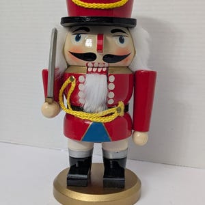 Puede incluir: Un cascanueces de madera con uniforme rojo y sombrero alto, sosteniendo una espada plateada. La figura tiene barba blanca, bigote negro y botas negras. La base es dorada.