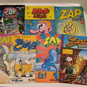 Op de afbeelding: Een verzameling van zeven Zap Comix-stripboeken met kleurrijke grafische illustraties en tekst op de covers. De uitgaven bevatten verschillende nummers en titels zoals Zap Comix nr. 0, nr. 1, nr. 2, nr. 3 en nr. 5, gerangschikt in een raster.