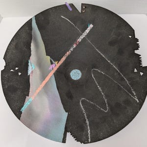 May include: A circular black wall plaque featuring a textured surface with a large, light-coloured triangular shape, a central blue circle, and white chalk-like lines. The edges are irregular with small cutouts and a small purple accent piece.