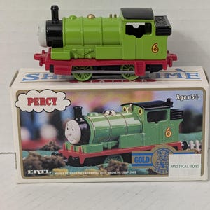 May include: A green die-cast metal toy train engine numbered 6, featuring a black roof, red chassis, and a white face. It sits atop its original rectangular cardboard packaging, which displays the name Percy, the Ertl logo, and the text Ages 3+.