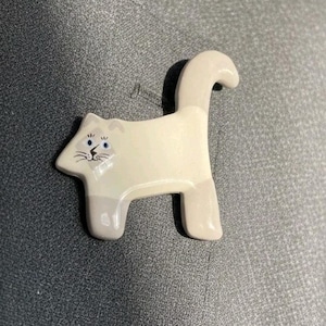 May include: A ceramic cat-shaped pin with a cream-colored body and gray accents. The cat has blue eyes and a whimsical design. The pin is attached to a silver pin backing.