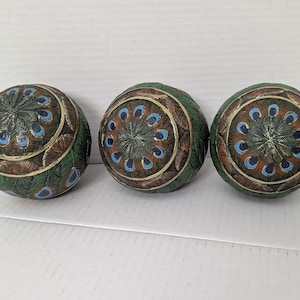 May include: Three spherical decorative objects featuring a detailed peacock feather pattern in shades of green, brown, and blue. Each sphere displays a circular arrangement of eye-like motifs radiating from the center, with textured surfaces.