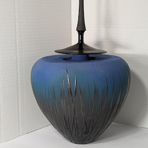 Puede incluir: Un jarrón decorativo azul y negro con una tapa negra alta y delgada. El jarrón tiene un degradado de azul a negro, con líneas verticales grabadas. La tapa es de un diseño oscuro y elegante.