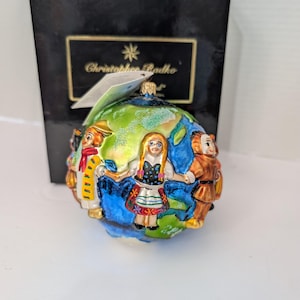 May include: A Christmas bauble with a globe design, featuring figures in traditional dress. The ornament has a glossy finish with blue, green, and gold accents. It is presented in a black box with the brand name "Christopher Radko".