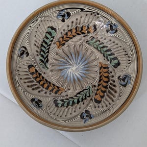May include: A round ceramic bowl featuring a cream-colored base with intricate slip-trailed patterns. The design includes a central blue starburst surrounded by alternating brown and green feather-like motifs, all set against fine black lines.