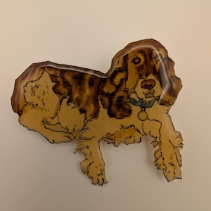 May include: A vintage-style dog-shaped brooch. The brooch features a light brown and tan color scheme with a detailed illustration of a dog. The dog is wearing a blue collar with a gold-colored tag. The brooch is likely made of enamel or a similar material.