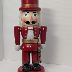 Puede incluir: Figura de cascanueces de madera roja con sombrero alto y banda dorada. La figura tiene barba y pelo blancos, ojos azules y bigote negro. La base es roja y los zapatos son negros. Mide aproximadamente 25 cm de altura.