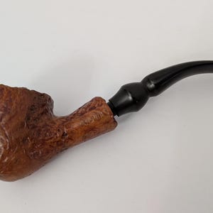 May include: A brown tobacco pipe with a textured bowl and a curved, black stem. The pipe is made of wood and has a classic design. The pipe is a smoking accessory.