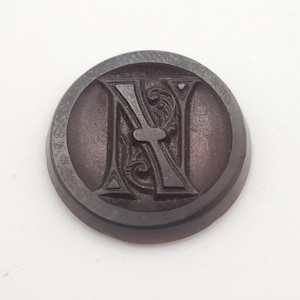 Könnte beinhalten: Ein runder, dunkelbrauner Knopf mit einem erhabenen, geprägten Großbuchstaben N in der Mitte. Der Buchstabe ist von komplizierten, wirbelnden Filigranmustern umgeben, die in die Oberfläche des Knopfes eingelassen sind und eine Textur erzeugen.