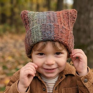 May include: A crocheted beanie with pointed cat-ear corners, featuring a variegated yarn pattern in blended shades of orange, brown, green, and blue. The hat has a textured, ribbed stitch construction and sits snugly on the head with a folded brim.