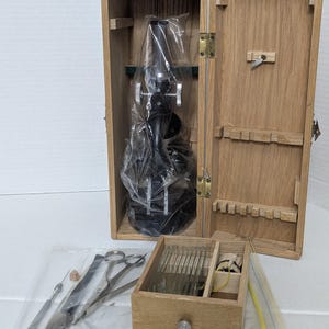 May include: A black metal microscope encased in a clear plastic bag inside a tall wooden storage box. Beside it sits a small wooden drawer containing glass slides and a plastic bag filled with metal dissection tools including scissors.