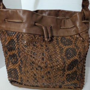 May include: A brown bucket bag featuring a textured snakeskin pattern on the lower body and a smooth leather upper section. The bag includes a drawstring closure with a leather cord, a single shoulder strap, and a flat base with small metal feet.
