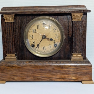 May include: Antique wooden mantel clock with a round clock face. The clock has a dark brown wooden frame with gold-colored accents on the columns and base. The clock face has black numerals and hands.