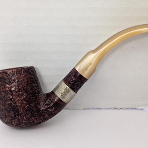 May include: A brown tobacco pipe with a textured bowl and a light-colored, curved stem. The pipe has a silver band with text near the stem. The bowl is a dark brown color, and the stem is a light tan color.