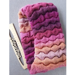 Könnte beinhalten: Handgestrickter Schal in Rosa-, Lila- und Pfirsichtönen mit einem wellenförmigen, strukturierten Muster. Der Schal ist gefaltet und zeigt den Farbverlauf. Ein Holzlineal mit dem Text "Astraelina" liegt daneben.