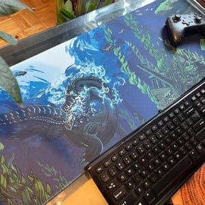 Può includere: Un tappetino per mouse da gioco blu e verde con un'illustrazione di un mostro marino nero. Il mostro è circondato da acqua blu e alghe verdi. Una tastiera nera e un controller di gioco nero sono sul tappetino per mouse.
