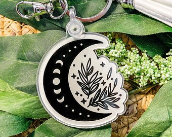 Könnte beinhalten: Ein schwarzer und weißer Halbmond-Schlüsselanhänger mit einem Blatt-Zweig-Design und einem weißen Quasten. Der Schlüsselanhänger ist aus klarem Acryl und hat einen silbernen Metallring.