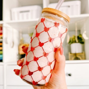 May include: A glass jar with a pink smoothie inside, decorated with a red heart-shaped net. The jar has a wooden lid and a straw sticking out of the top.