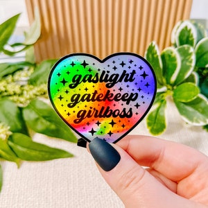Peut inclure: Un autocollant en forme de cœur holographique arc-en-ciel avec le texte "gaslight gatekeep girlboss" en noir.