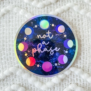 Può includere: Un adesivo rotondo con finitura olografica che presenta uno sfondo di galassia con un cerchio di fasi lunari e il testo "not a phase" in bianco.