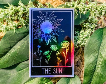 Puede incluir: Una pegatina negra y holográfica iridiscente con un dibujo de línea del sol y flores. El texto "THE SUN" está impreso en la parte inferior de la pegatina.