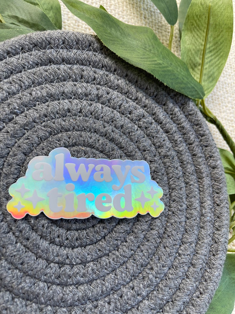 Op de afbeelding: Een holografische sticker met de tekst "always tired" in een wolkvorm met sterren aan beide kanten.