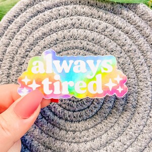 Op de afbeelding: Een holografische sticker met een regenboog-tie-dye achtergrond en witte tekst die "always tired" zegt met twee witte sterren aan beide kanten.