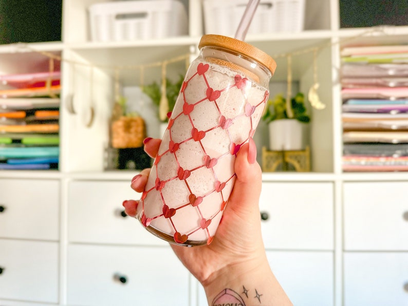 May include: A pink smoothie in a clear glass with a red heart pattern. The glass has a bamboo lid and a straw.