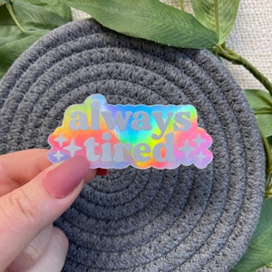 Op de afbeelding: Een holografische sticker met een regenboog-tie-dye achtergrond en de tekst "always tired" in wit. De sticker heeft twee kleine witte sterren aan weerszijden van de tekst.