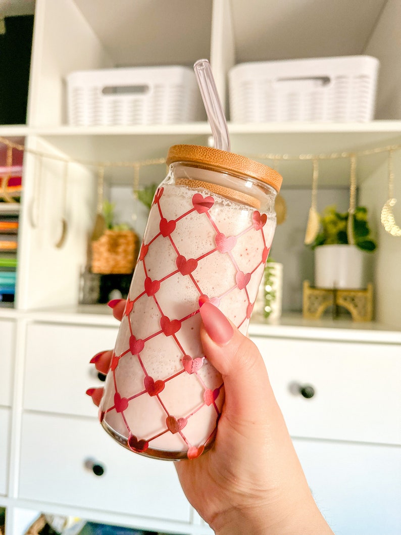 May include: A glass jar with a pink smoothie and a clear straw. The jar has a wooden lid and a pink and white heart pattern.