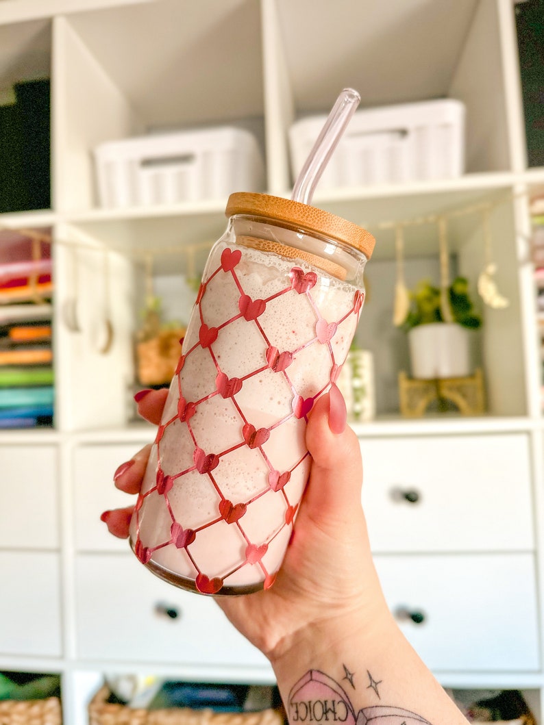 May include: A glass jar with a pink smoothie and a clear straw. The jar has a wooden lid and is decorated with a red heart pattern.