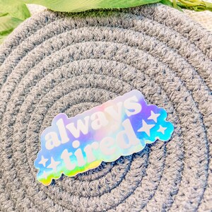 Op de afbeelding: Een holografische sticker met de tekst "always tired" in wit met een regenboogkleurige, iriserende achtergrond. De sticker heeft twee witte sterren aan de rechterkant.