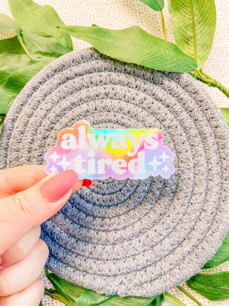 Op de afbeelding: Een holografische sticker met een regenboog-tie-dye achtergrond en witte tekst die "always tired" luidt met twee witte sterren aan weerszijden van de tekst.