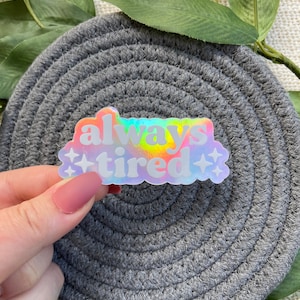 Op de afbeelding: Een holografische sticker met de tekst "always tired" in een regenboogkleurenschema. De sticker heeft een wolkvorm met twee kleine sterren aan weerszijden van de tekst.