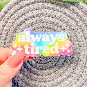 Op de afbeelding: Een holografische sticker met een regenboog-tie-dye achtergrond en de tekst "always tired" in wit. De sticker heeft twee kleine witte sterren aan weerszijden van de tekst.