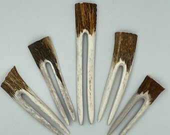 May include: Five U-shaped hair forks crafted from natural antler material. Each piece features a dark brown, textured top section that transitions into two smooth, light-colored, tapered prongs. The forks are arranged in a fan-like formation.