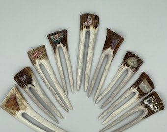 May include: A collection of nine U-shaped hair forks crafted from textured brown and white antler material. Each fork features a unique, polished gemstone inlay at the top, showcasing various colors and patterns including pink, green, and clear stones.