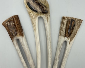 May include: Three hair forks crafted from textured, light-colored antler material. Each fork features a two-pronged design and is inlaid with a smooth, polished, dark brown and tan stone accent at the top, showcasing natural patterns and variations.