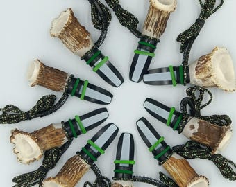 May include: A collection of hunting calls made from deer antlers, black and white plastic, and green rubber bands. Each call has a black and green cord for wearing around the neck. The calls are arranged in a circular pattern against a white background.