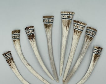 May include: A collection of seven curved, tapered bone-like objects arranged in a fan shape. Each piece features a smooth, off-white surface with brown, weathered-looking textures at the wider end, accented by a dark, intricate horizontal band.