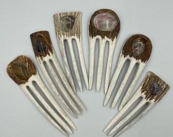 May include: Six hair forks crafted from textured brown and white antler material, each featuring a unique polished gemstone embedded at the top. The forks have three prongs and vary in shape, showcasing natural patterns and distinct mineral colors.