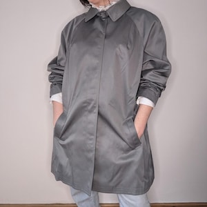 May include: A grey, knee-length raincoat with a button closure and two front pockets. The coat has a collared neckline and long sleeves. A white shirt is visible underneath. The coat is made of a smooth, shiny material.