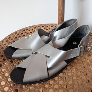 May include: A pair of silver sandals featuring wide crisscross straps over the top, an open toe design, and a black footbed. The sandals include a side strap with a metal buckle closure and a white circular label with the letter M on the heel.