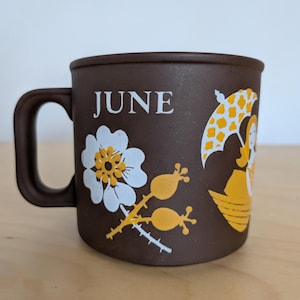 May include: A brown ceramic mug featuring the word JUNE in white text. The design includes a white and yellow flower illustration next to a yellow graphic of a person holding a patterned umbrella while sitting in a small boat on the water.