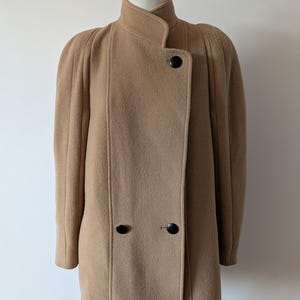 以下が含まれることがあります： ハイカラー、縦のプリーツが入った構築的なショルダー、3つの大きな丸い黒いボタンで留めるフロントクロージャーが特徴のタンカラーのウール混コート。マネキンに飾られた、ストレートなシルエットと長袖のコート。