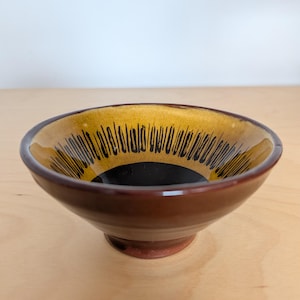 May include: A ceramic bowl with a glossy dark brown exterior and a footed base. The interior features a deep black center surrounded by a wide band of mustard yellow glaze, decorated with a series of vertical black brushstroke lines.
