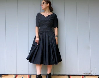 Può includere: Una donna che indossa un abito nero con un corpetto aderente e una gonna ampia. Indossa stivaletti neri alla caviglia e ha i capelli tirati indietro.