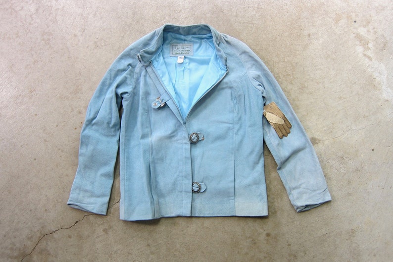 May include: A light blue suede jacket with a zip fastening and three metal buckles. The jacket has a label that says "L.A. Look" on it.