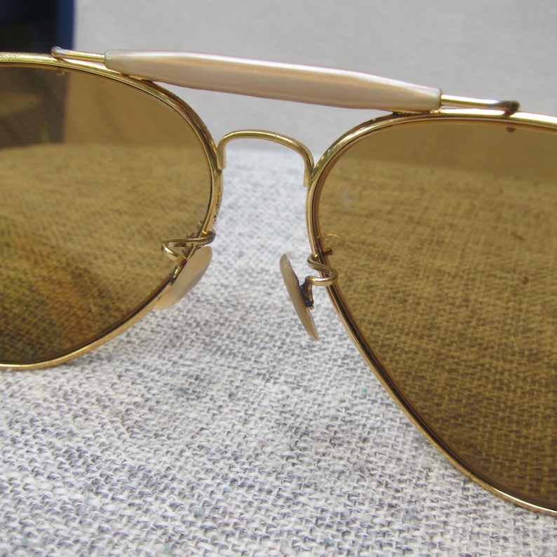 Puede incluir: Primer plano de gafas de sol de aviador con montura dorada y lentes marrones. Las gafas de sol presentan un puente y almohadillas nasales de color claro. Las lentes son de color &aacute;mbar c&aacute;lido. La montura es de estilo aviador cl&aacute;sico.